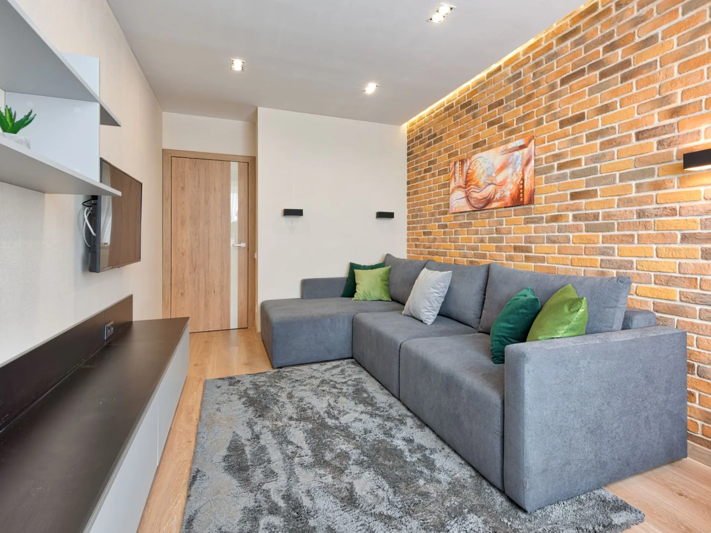 A grey sectional sofa with green cushions beside an exposed brick wall.
