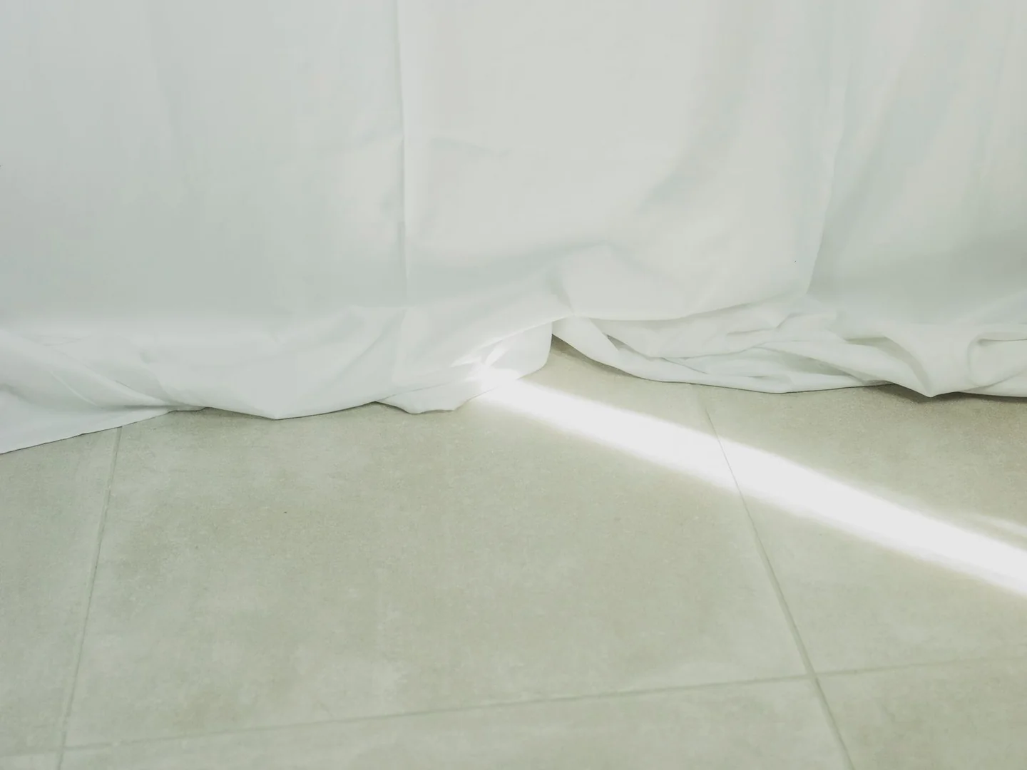 White curtain hems resting in soft folds on a pale tiled floor.