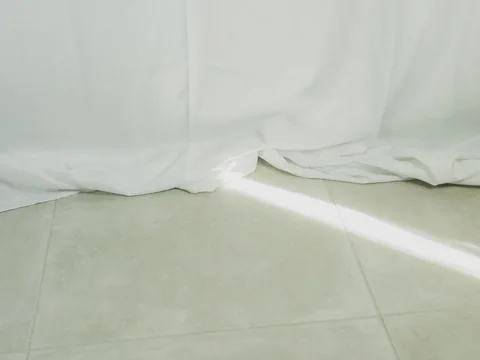 White curtain hems resting in soft folds on a pale tiled floor.